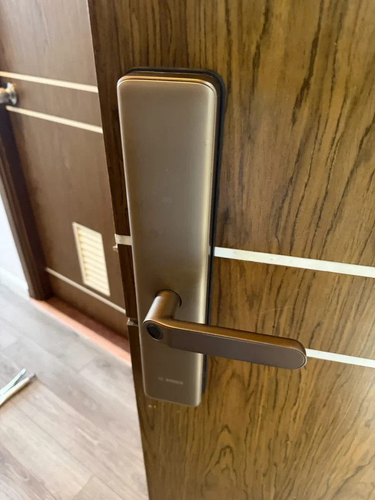 Khóa điện tử màu đồng đang lắp trên cửa gỗ, băng dính đánh dấu vị trí khoan